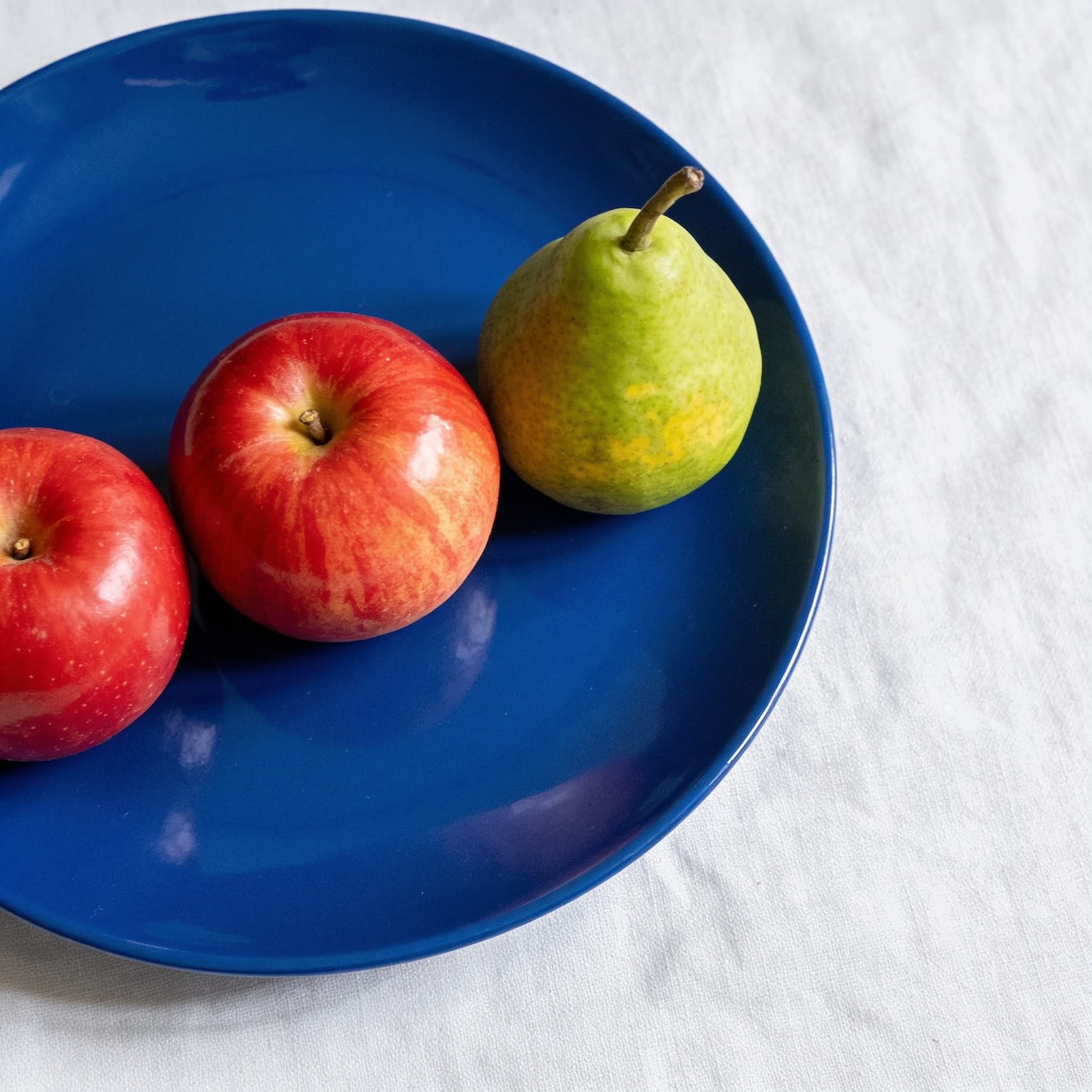 Seedream 4.5: Three red apples and one green pear arranged in a single row on a blue plate, top-down view, white tablecloth