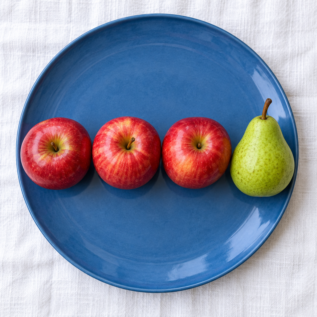 GPT-Image 2: Three red apples and one green pear arranged in a single row on a blue plate, top-down view, white tablecloth