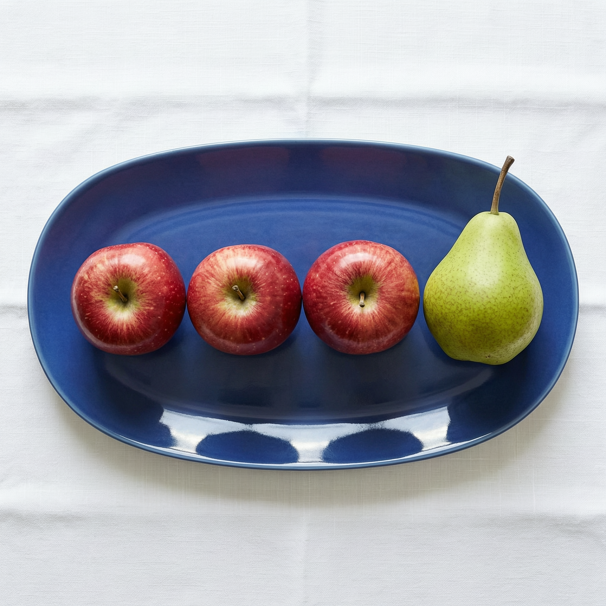 WAN 2.7 Pro: Three red apples and one green pear arranged in a single row on a blue plate, top-down view, white tablecloth