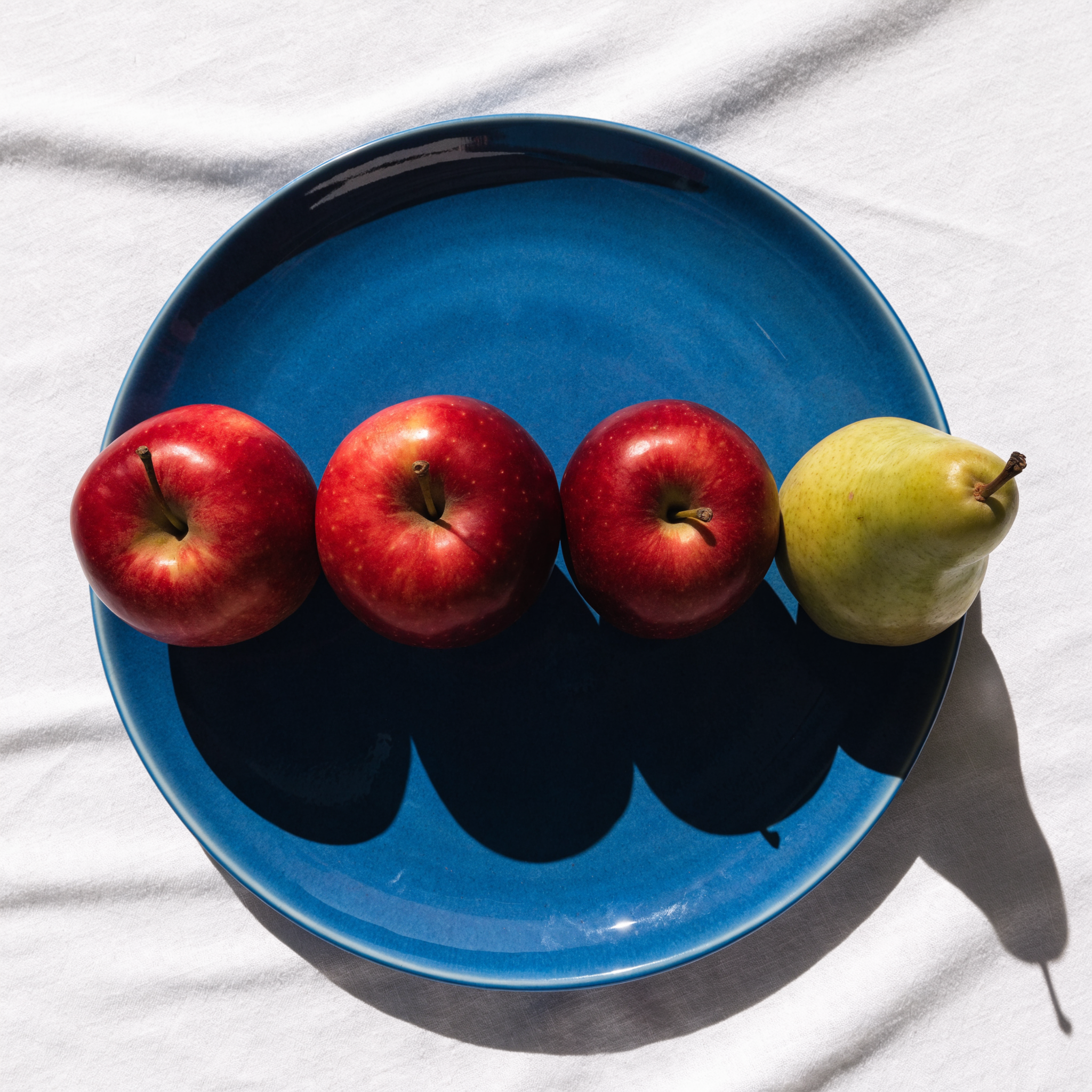 Recraft V4.1 Pro: Three red apples and one green pear arranged in a single row on a blue plate, top-down view, white tablecloth